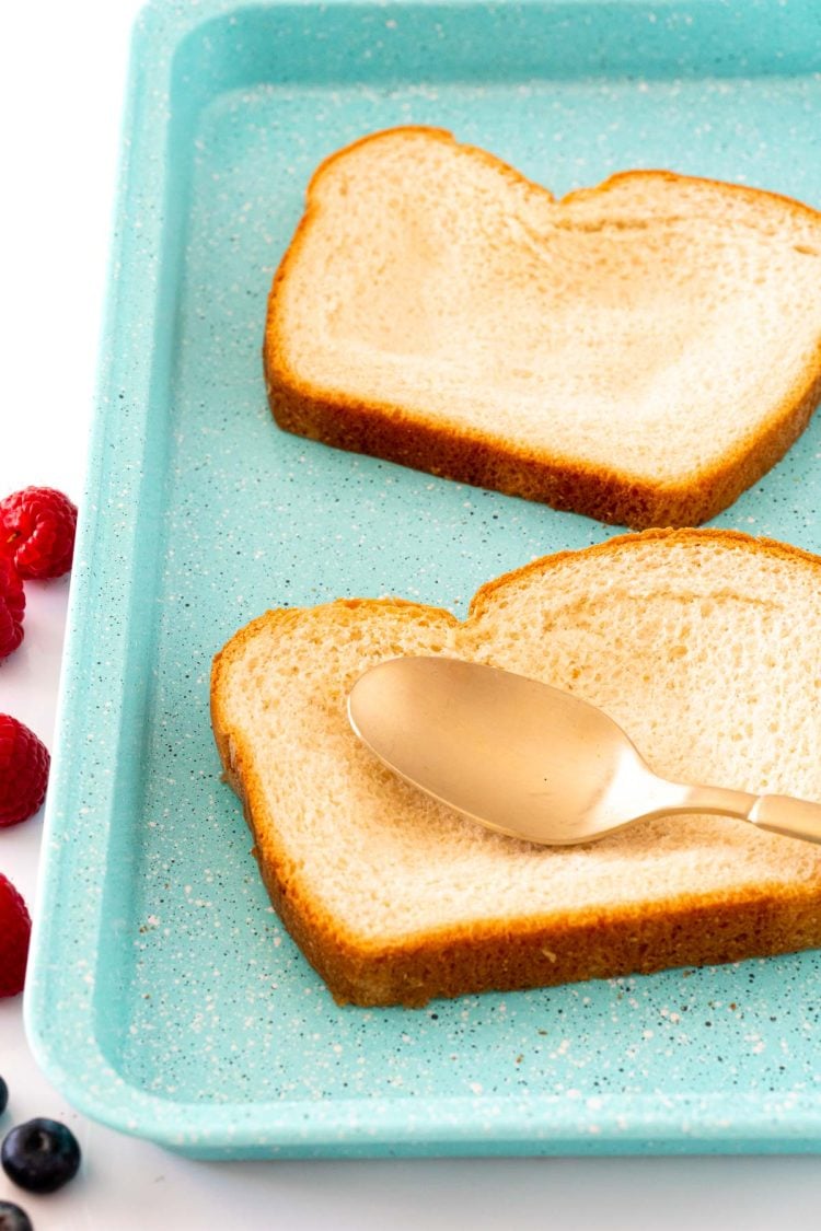 Two slices of toast on a blue baking sheet with a spoon smooshing down the centers of the bread to create a well.