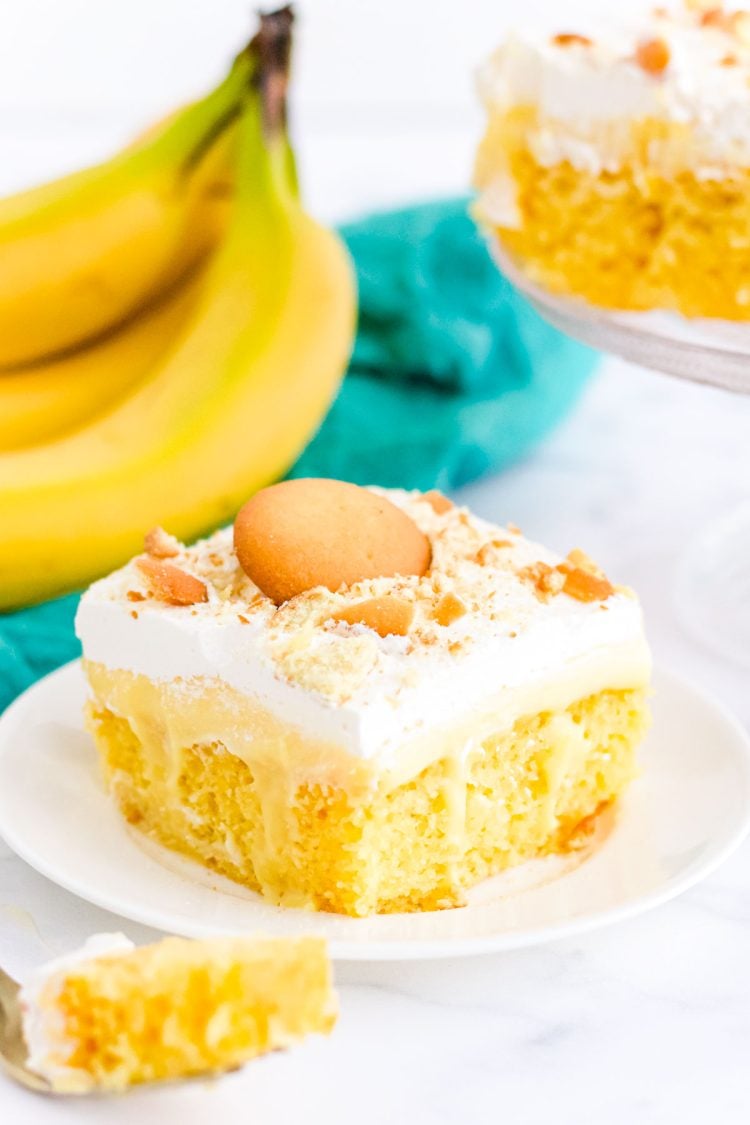 A slice of banana cream poke cake on a white plate with a teal napkin behind it.