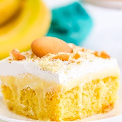 Close up of a slice of banana poke cake on a white plate with bananas in the background.