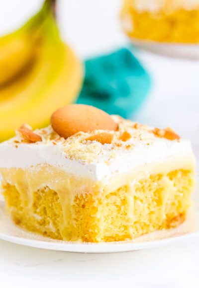 Close up of a slice of banana poke cake on a white plate with bananas in the background.