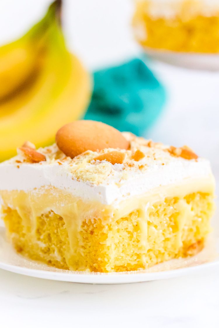 Close up of a slice of banana poke cake on a white plate with bananas in the background.