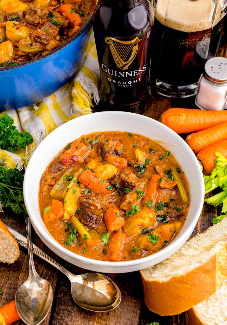 A white bowl with beef stew on a wooden table with ingredients, spoons, and Guinness around it.