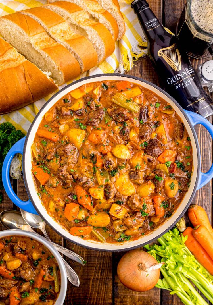 Overhead photo of a Dutch oven filled with Irish beef stew on a wooden table with Guinness and bread around it.
