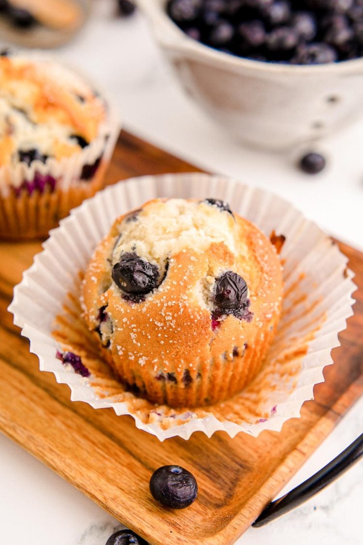 A blueberry muffin on a wooden tray with the liner being peeled off.