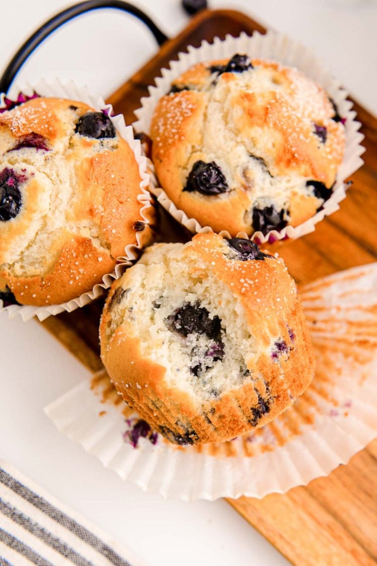 Three blueberry muffins on a wooden tray, one is missing a bite.