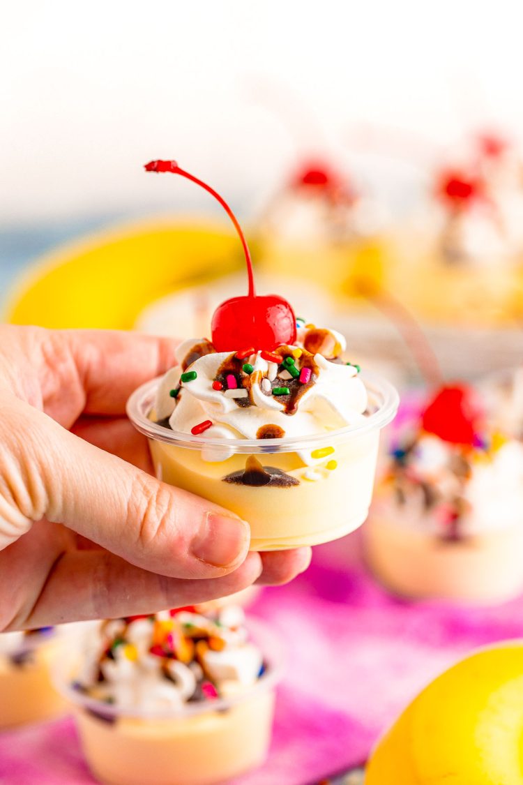 A woman's hand holding banana split pudding shots to the camera.