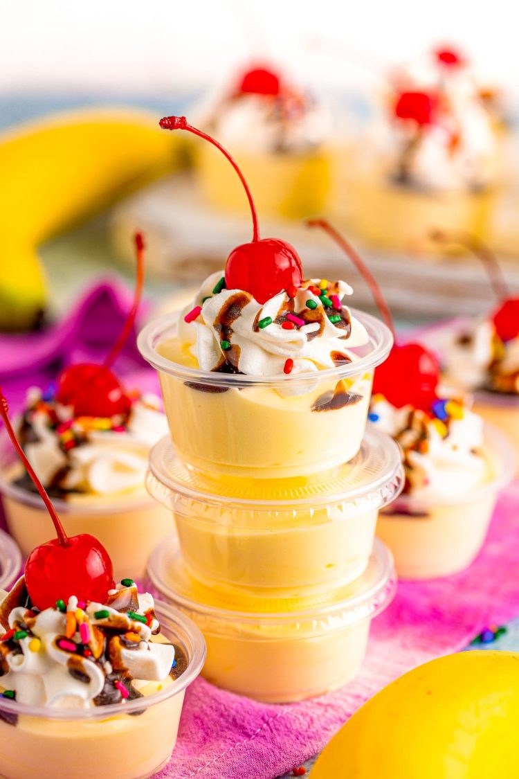 A stack of three banana split pudding shots topped with whipped cream, chocolate syrup, and a cherry.