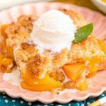 Close up photo of a slice of peach cobbler on a pink plate on a blue napkin.