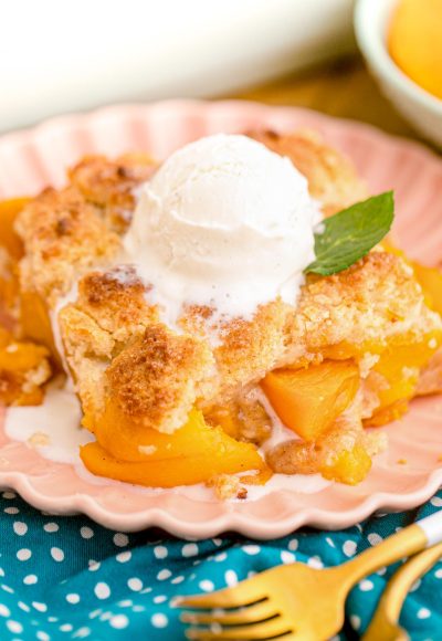 Close up photo of a slice of peach cobbler on a pink plate on a blue napkin.