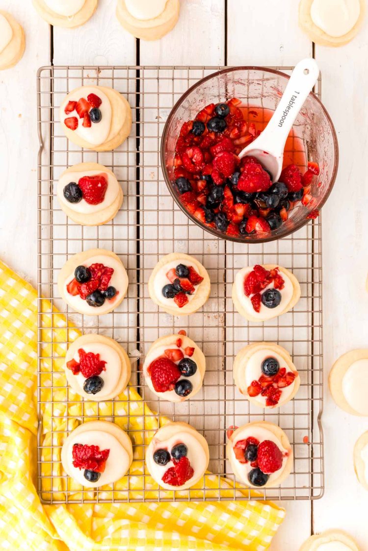 Overhead photo of shortcake cookies topped with frosting and berries on a wire rack with a bowl of macerated berries next to them.