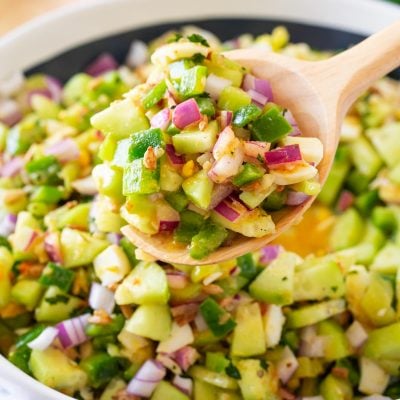 A serving spoon scooping cucumber salad out of a white serving bowl.