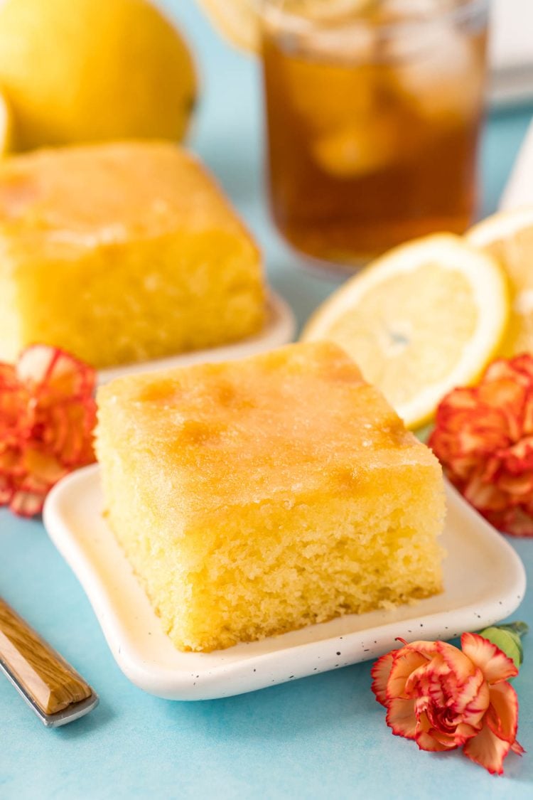 A slice of lemon drizzle cake on a white plate on a blue surface.