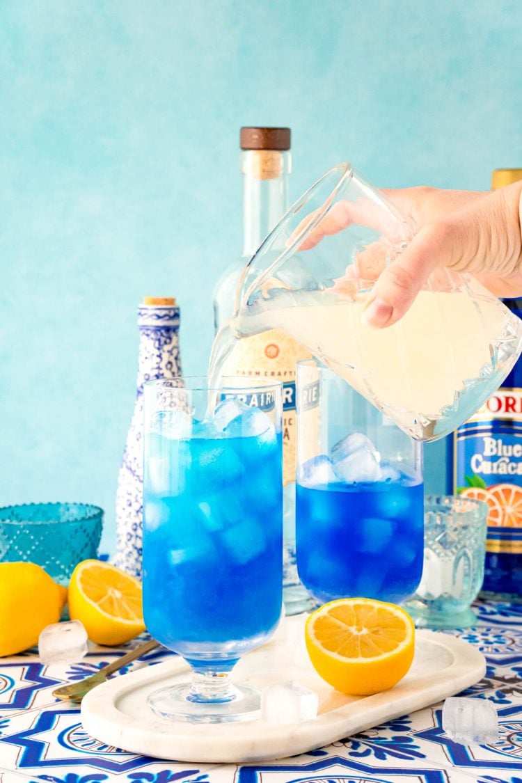 Lemonade being poured from a glass pitcher into a glass to make a blue cocktail.