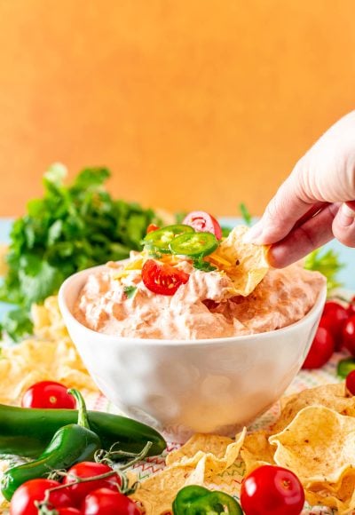 Close up photo of a woman's hand scooping boat dip out of a white bowl with a tortilla chip.