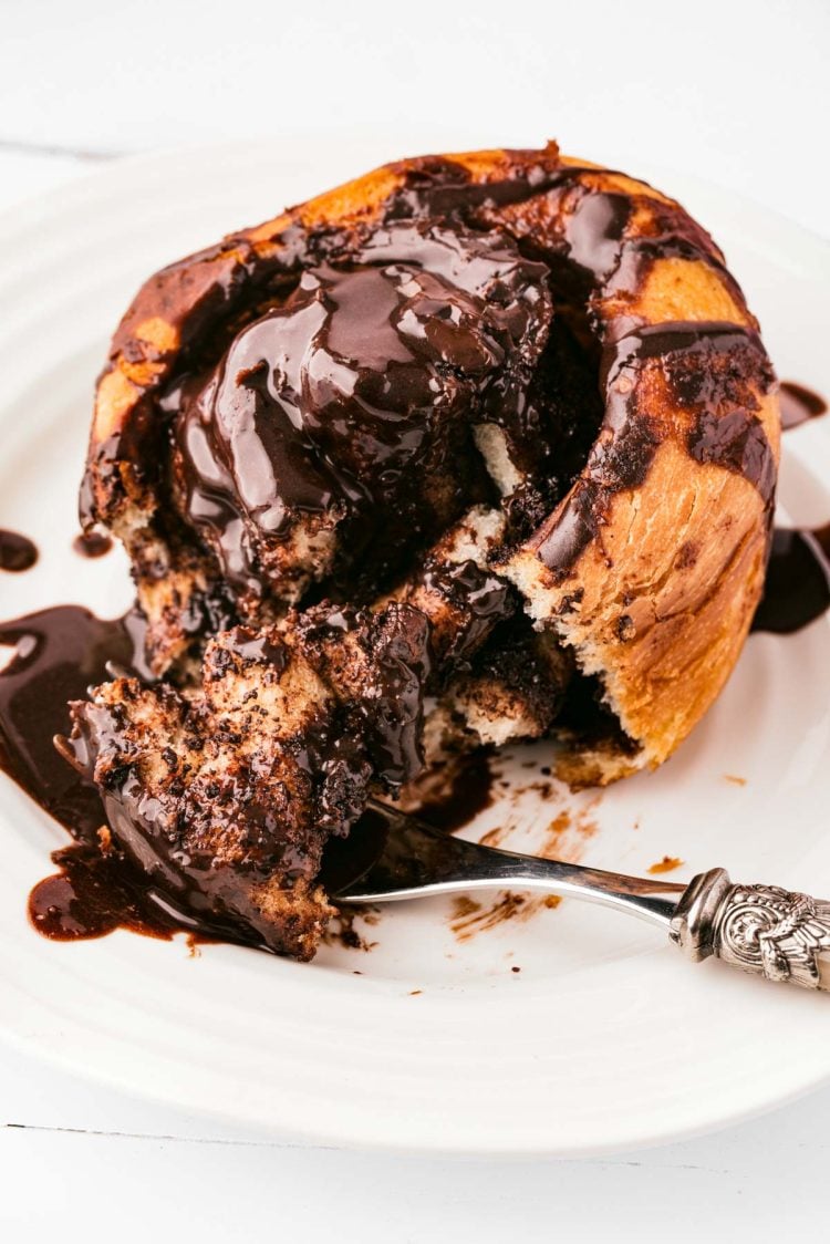 A chocolate cinnamon roll on a white plate with a fork taking a bite out of it.