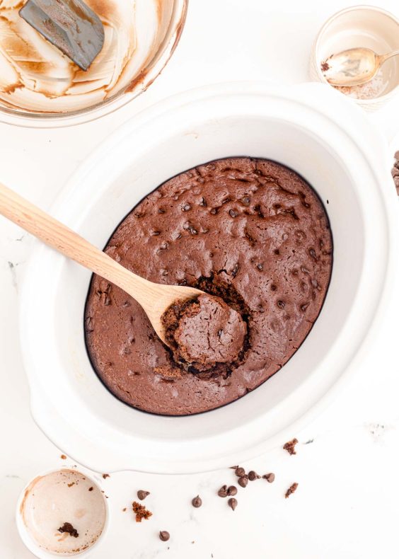 A wooden spoon scooping chocolate cake out of a white crockpot.