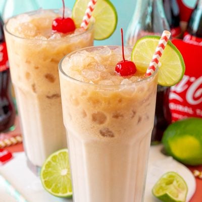 Two glasses with dirty sodas in them on a white serving board with coca cola in the background.