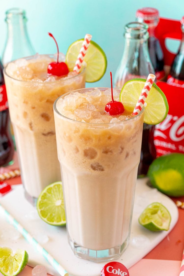 Two glasses with dirty sodas in them on a white serving board with coca cola in the background.