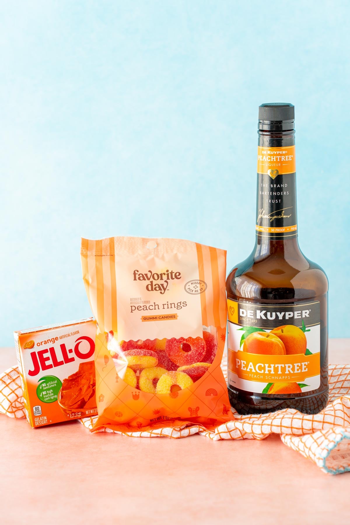 A box of Orange jello, a bag of peach rings, and a bottle of peach schnapps on a tan surface.