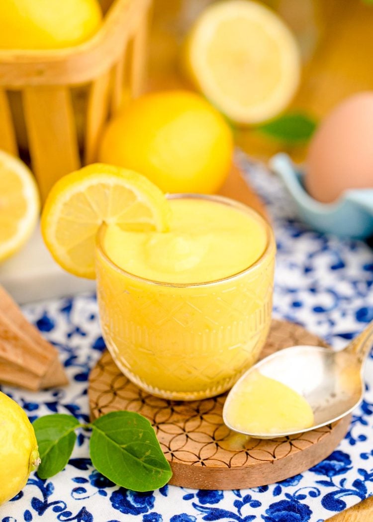 Close up photo of lemon curd in a glass cup on a wooden coaster on a blue and white napkin.