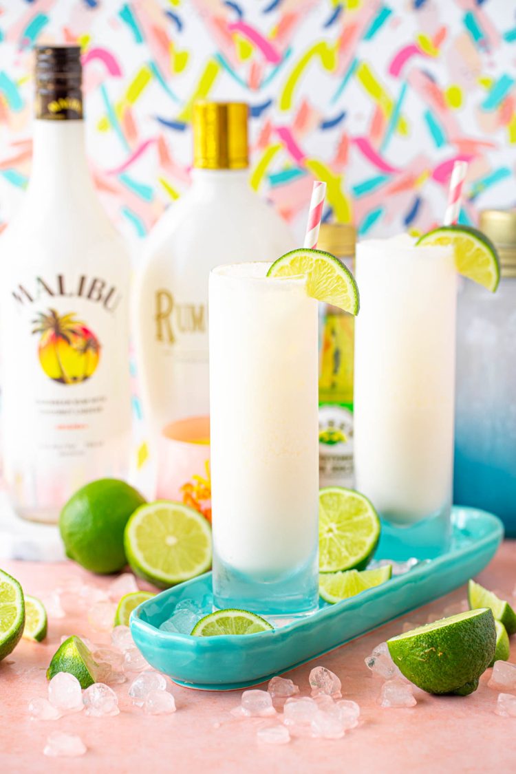 Two glass with white cocktail on a teal dish surrounded by limes and ice.