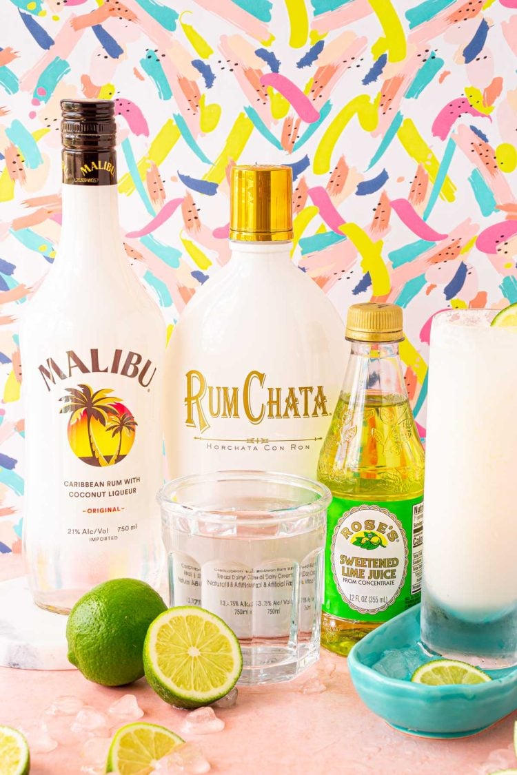 Malibu, RumChata, Lime Juice bottles and a glass of club soda on a pink surface surrounded by limes.
