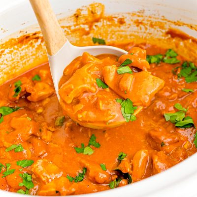 A ladle scooping slow cooker butter chicken from a white crockpot.