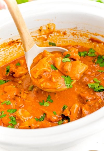 A ladle scooping slow cooker butter chicken from a white crockpot.