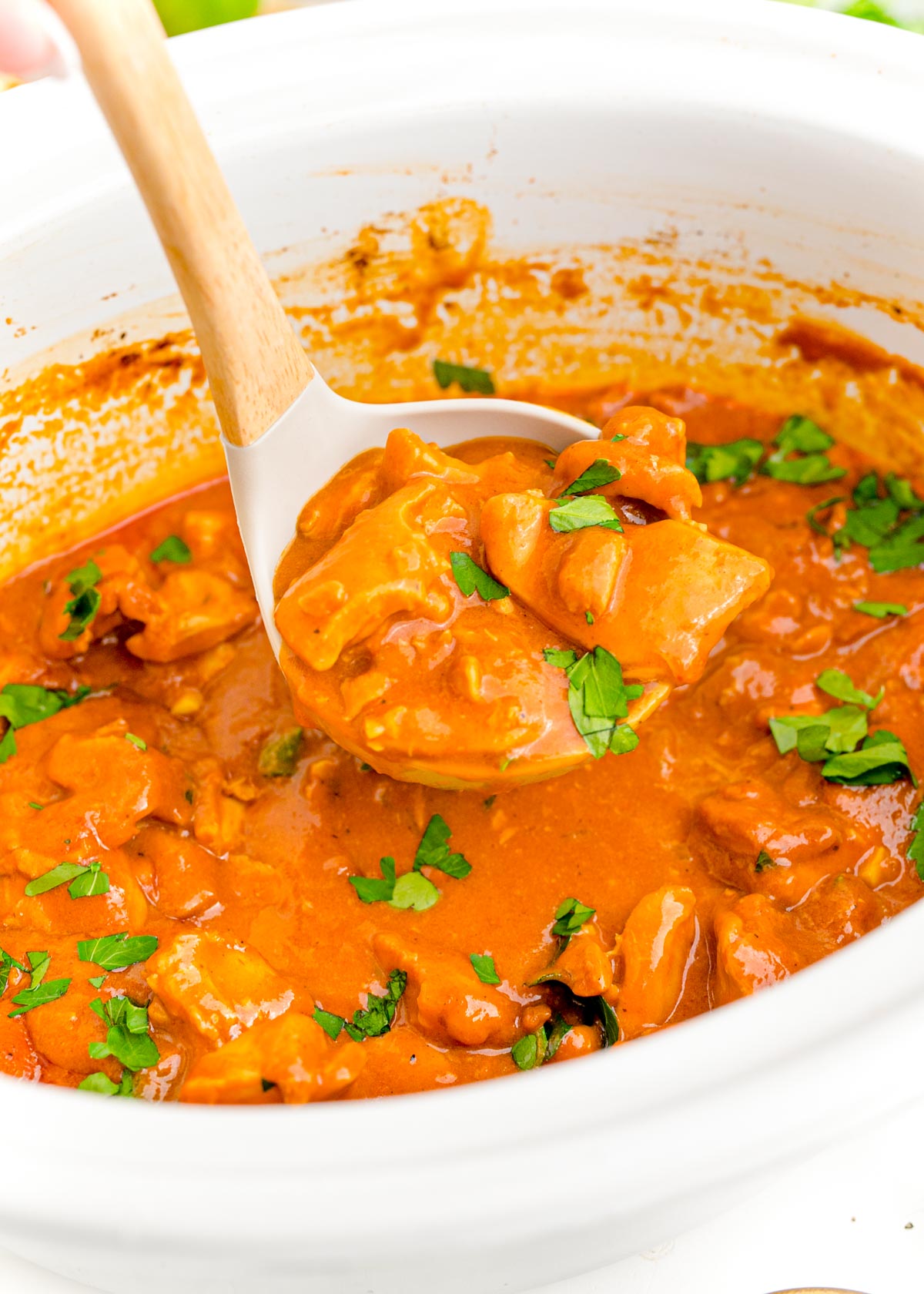 A ladle scooping slow cooker butter chicken from a white crockpot.