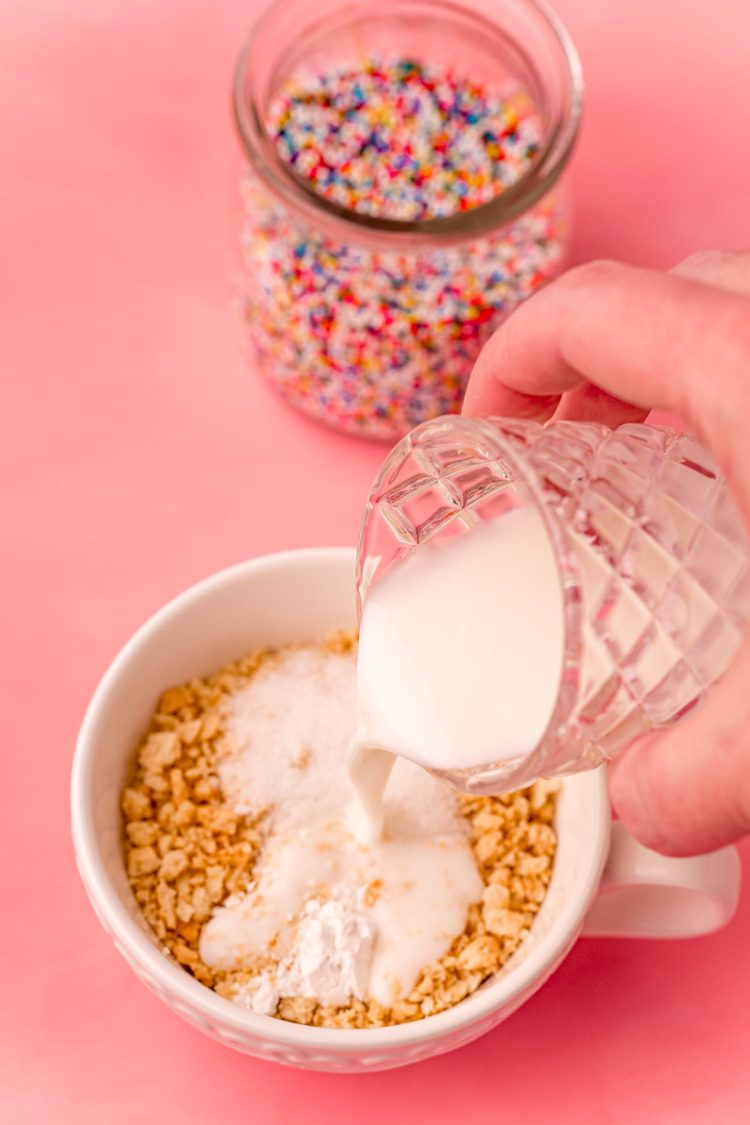 A woman's hand pouring milk into a white tea cup with crushed oreos on a pink surface.
