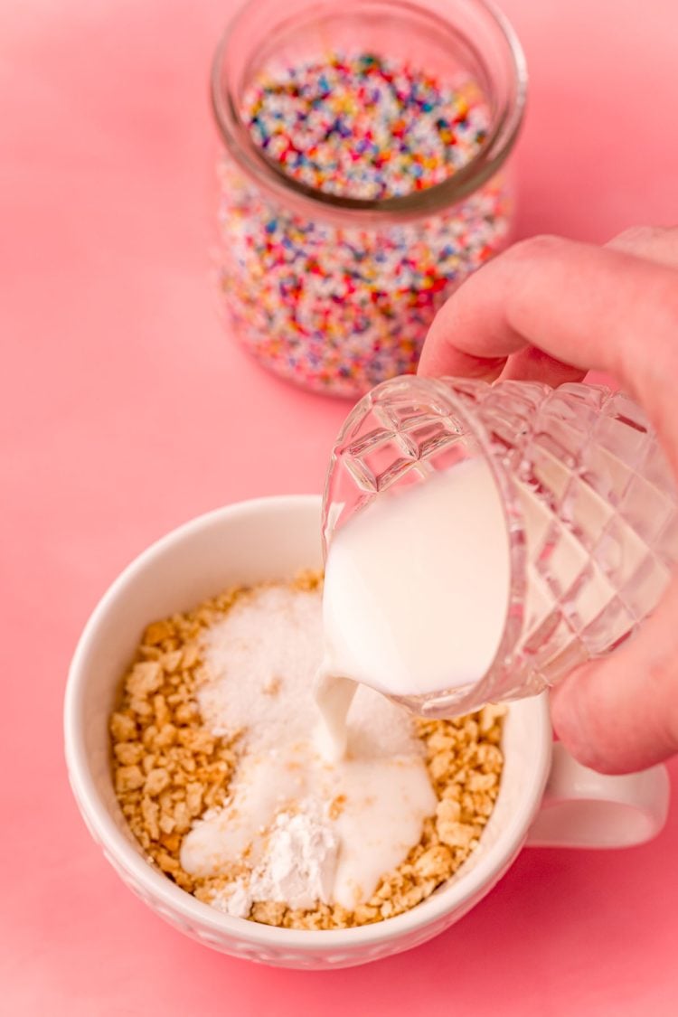 A woman's hand pouring milk into a white tea cup with crushed oreos on a pink surface.