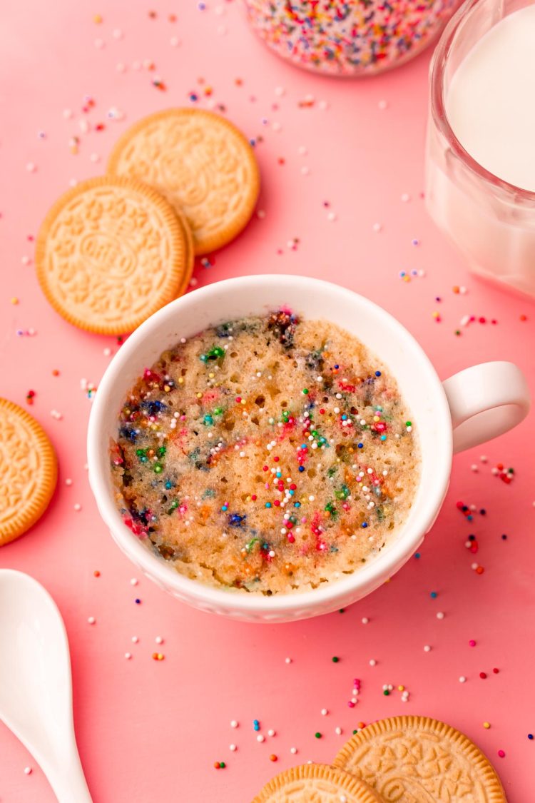 A funfetti mug cake in a white cup on a pink surface.