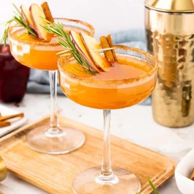 Two coupe glasses filled with an apple cider bourbon cocktail on a cutting board.