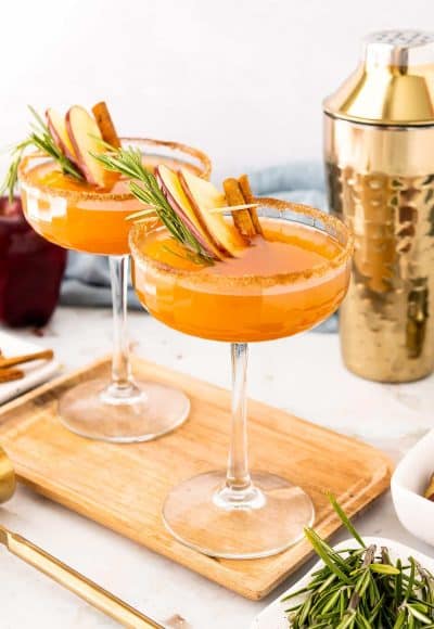 Two coupe glasses filled with an apple cider bourbon cocktail on a cutting board.