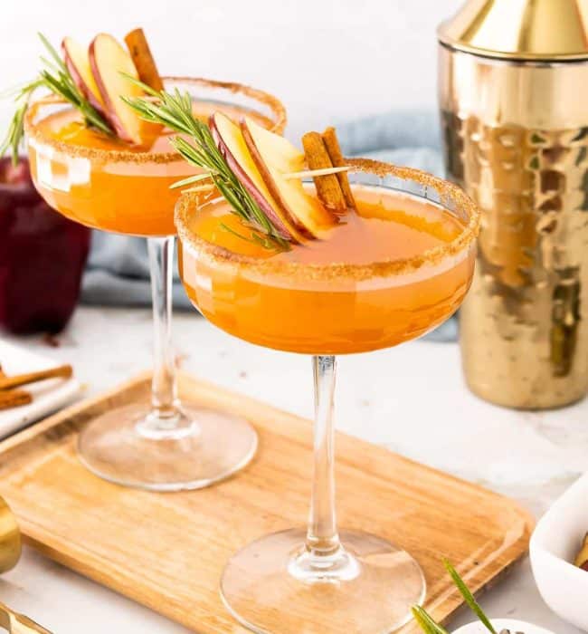 Two coupe glasses filled with an apple cider bourbon cocktail on a cutting board.