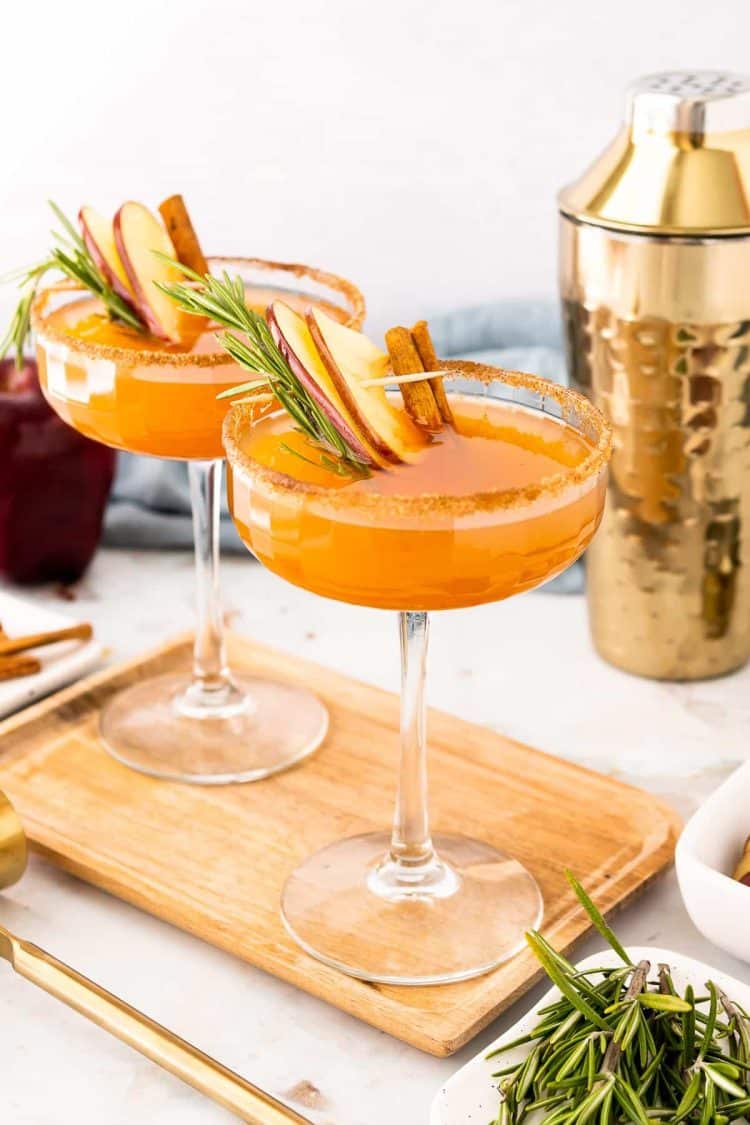 Two coupe glasses filled with an apple cider bourbon cocktail on a cutting board.