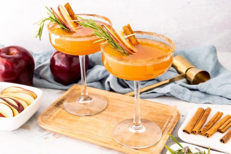Two coupe glasses filled with an apple cider bourbon cocktail on a cutting board.
