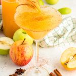 Apple cider martini surrounded by apples, cinnamon sticks, and a bottle of cider.