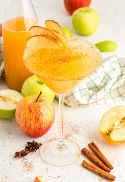 Apple cider martini surrounded by apples, cinnamon sticks, and a bottle of cider.