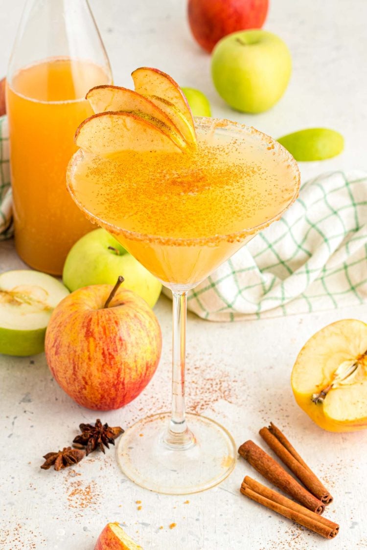 Apple cider martini surrounded by apples, cinnamon sticks, and a bottle of cider.