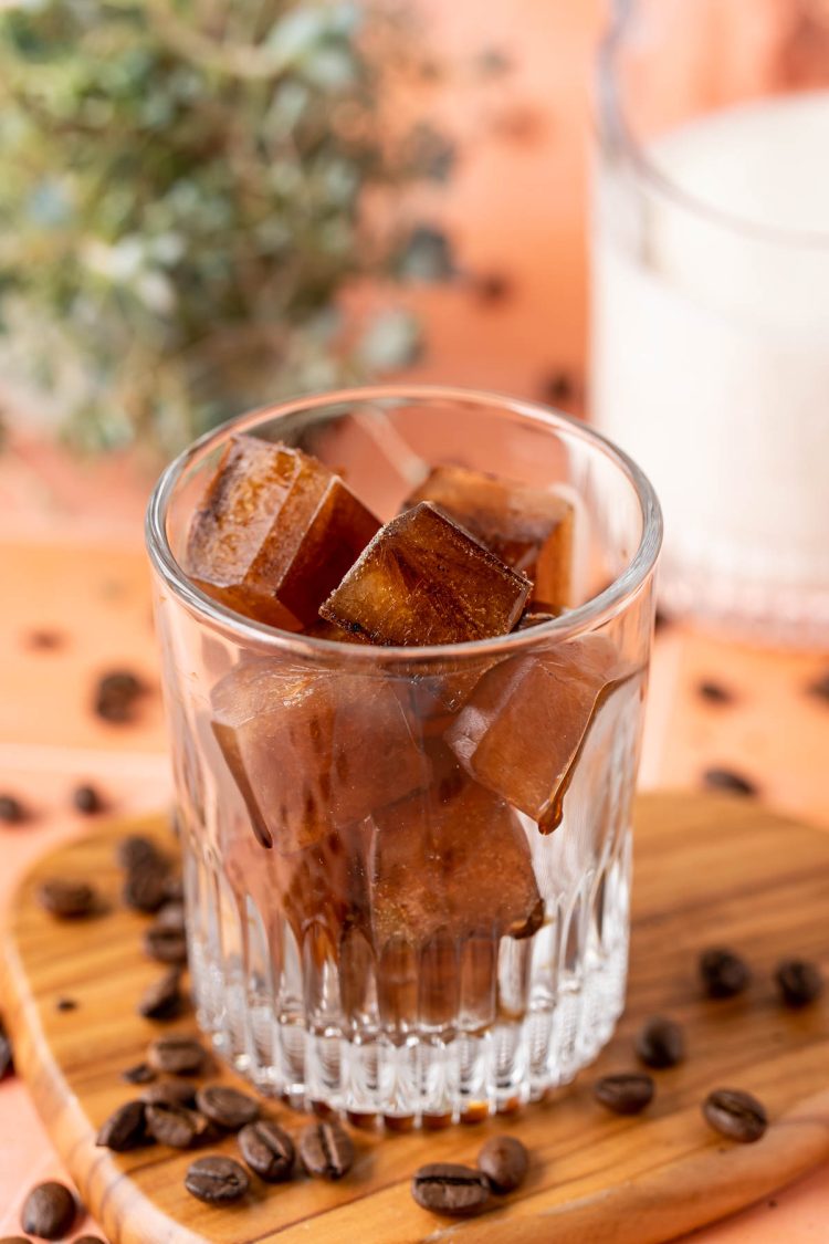 Coffee ice cubes in a glass with coffee beans scattered around it.