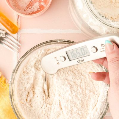 flour in a glass bowl with a woman's hand taking it's temperature with a kitchen thermometer.