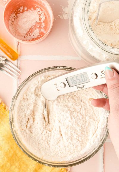 flour in a glass bowl with a woman's hand taking it's temperature with a kitchen thermometer.