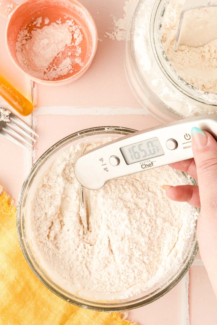 flour in a glass bowl with a woman's hand taking it's temperature with a kitchen thermometer.