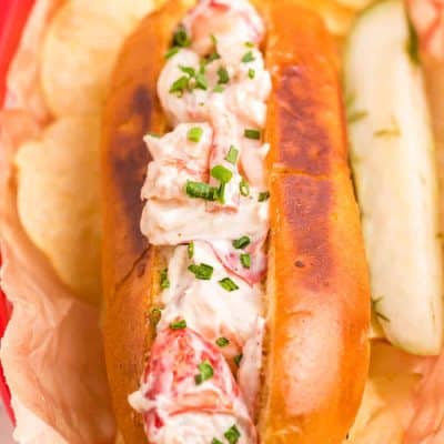 Close up photo of a lobster roll in a red basket with chips and a pickle.