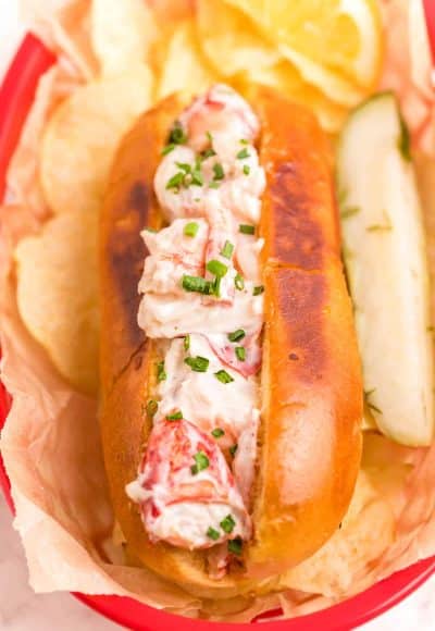 Close up photo of a lobster roll in a red basket with chips and a pickle.