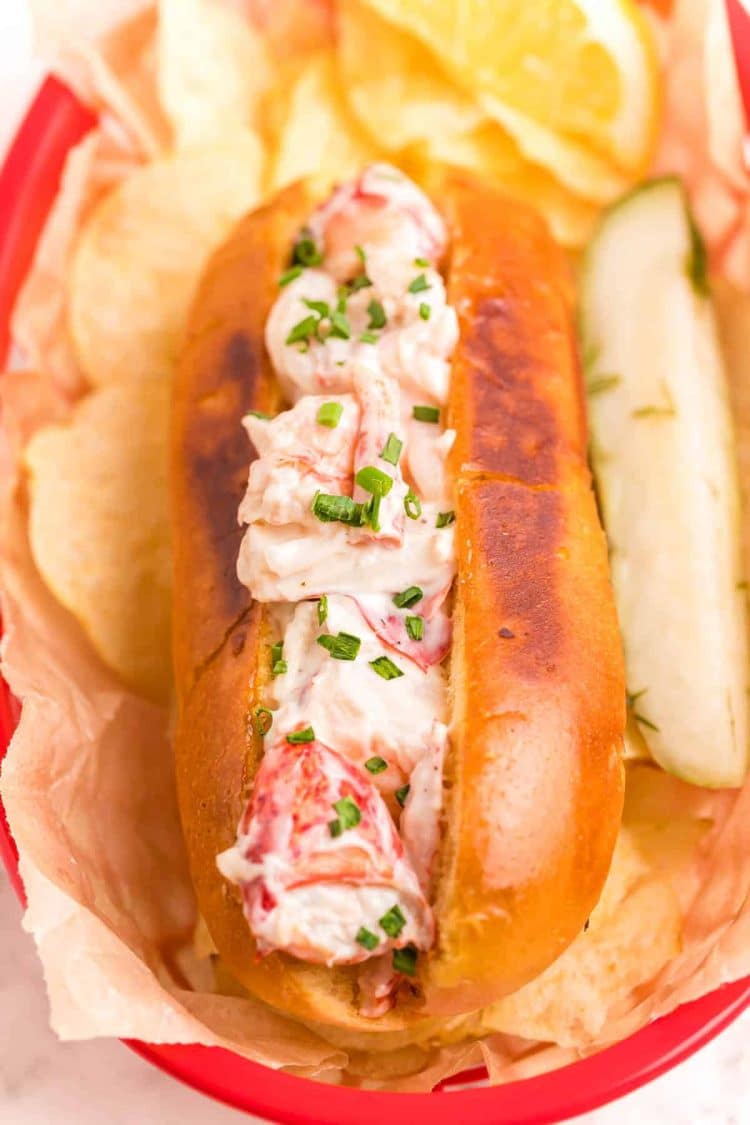 Close up photo of a lobster roll in a red basket with chips and a pickle.