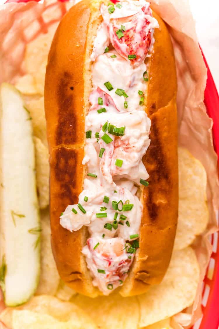 Close up photo of a lobster roll in a red basket with chips and a pickle.