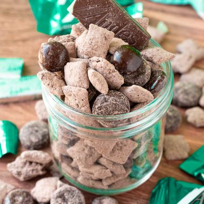 Mint chocolate muddy buddies in a jar on a wooden table surrounded my muddy buddies and mint candies.