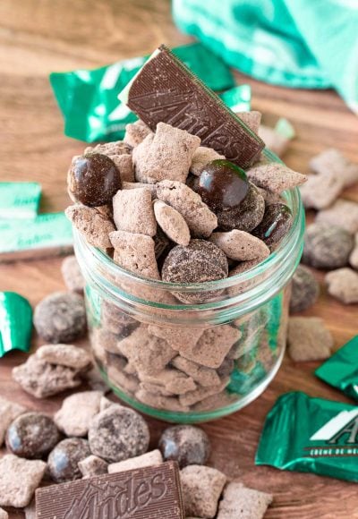 Mint chocolate muddy buddies in a jar on a wooden table surrounded my muddy buddies and mint candies.